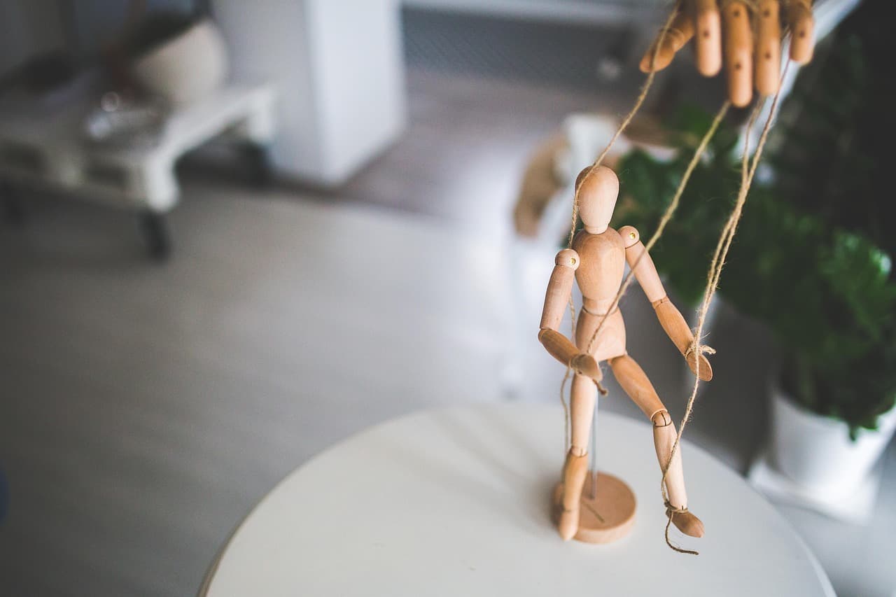 An articulated wooden mannequin standing on a white table, its strings held by a human hand above it