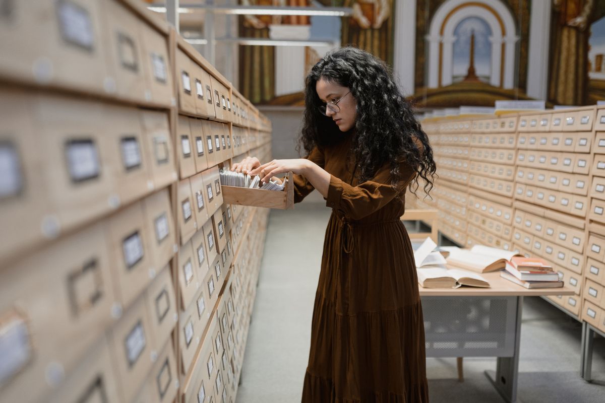 A reader in an archive room, going through a drawer of index cards by hand, open books on the table behind her.
